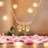 In the foreground, pink rose petals are scattered beneath a delicate Boy and Girl Children Pendant Hearth Necklace, crafted in 14k gold. Its three figure-shaped pendants glisten against soft, warm lights, creating a romantic ambiance—an ideal gift for Mother’s Day.